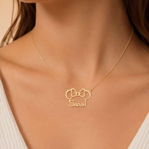 May include: A gold-toned necklace featuring a pendant shaped like a stylized mouse head silhouette with a bow at the top. The name Sarah is integrated into the lower portion of the design in a cursive script font, suspended from a delicate chain.