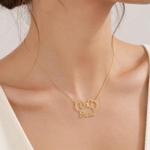 May include: A gold-toned necklace featuring a pendant shaped like a stylized mouse head with a bow. The name SARAH is integrated into the lower portion of the pendant design. The pendant is suspended from a delicate, thin gold-toned chain.