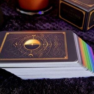 May include: A deck of rectangular cards with a black back featuring a gold celestial design of a sun and moon. The edges of the cards are colored in a vibrant rainbow gradient. The deck is positioned horizontally with the top card face up.