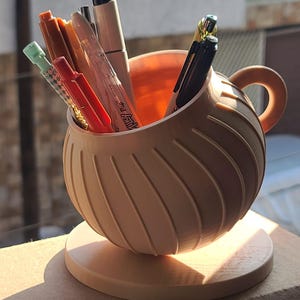 Puede incluir: Un portalápices esférico de color melocotón claro con un asa curva y textura acanalada diagonal, apoyado sobre una base circular. El soporte contiene varios bolígrafos, incluyendo instrumentos de escritura rojos, negros y metálicos.