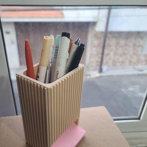 Puede incluir: Un portalápices rectangular de color beige con detalles acanalados verticales en el exterior. El soporte contiene varios bolígrafos y rotuladores de diversos colores, incluyendo naranja, blanco y negro. Una pequeña pila de notas adhesivas rosa claro está en la base.