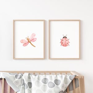 Op de afbeelding: Twee ingelijste wandkunstprints met aquarelillustraties. De linkerprint toont een roze libel met delicate vleugels, terwijl de rechterprint een roze lieveheersbeestje met zwarte stippen toont. Beide prints zitten in lichthouten lijsten.