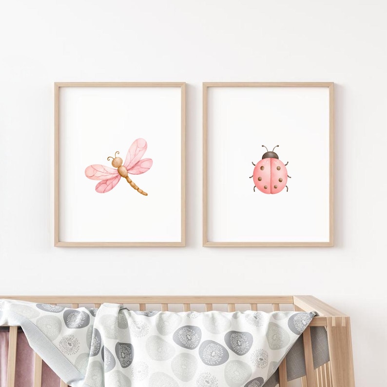 Op de afbeelding: Twee ingelijste wandkunstprints met aquarelillustraties. De linkerprint toont een roze libel met delicate vleugels, terwijl de rechterprint een roze lieveheersbeestje met zwarte stippen toont. Beide prints zitten in lichthouten lijsten.