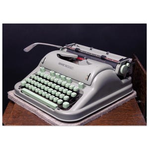 May include: A vintage grey Hermes 3000 typewriter with mint green keys. The typewriter is on a light grey surface, against a black background. The keys are in a QWERTY layout. It has a chrome carriage return lever. The model name is visible.