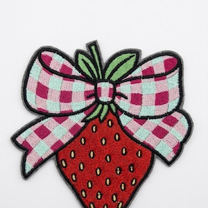 May include: An embroidered patch featuring a red strawberry with yellow seeds, topped with green leaves and a large bow with a pink and light blue checkered pattern. The entire design is outlined in a dark gray border against a plain background.