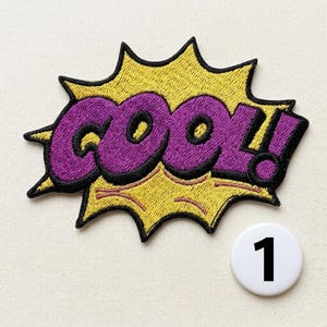 Op de afbeelding: Een geborduurde patch met het woord "cool!" in paars garen met een dikke zwarte omlijning, geplaatst op een gele tekstballon in stripstijl met zwarte randen. Een klein wit rond label met het zwarte cijfer 1 bevindt zich ernaast.