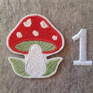 Puede incluir: Un parche bordado con forma de seta con un sombrero rojo con puntos blancos, una parte inferior verde claro y un tallo blanco. Dos hojas de color verde claro se encuentran en la base del tallo. El número 1 está bordado en blanco a la derecha.