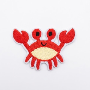 May include: An embroidered iron-on patch shaped like a crab. The crab is bright red with a cream-colored belly, two raised eyes with black pupils, and two large claws extended outward. It features a simple black stitched smile and a white border.