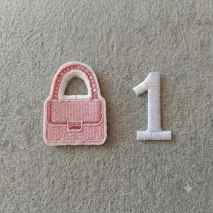 Op de afbeelding: Een roze geborduurde patch in de vorm van een handtas met een witte rand en een aparte witte geborduurde patch in de vorm van het cijfer één. Beide patches zijn voorzien van gedetailleerd stikwerk op een grijze, getextureerde ondergrond.