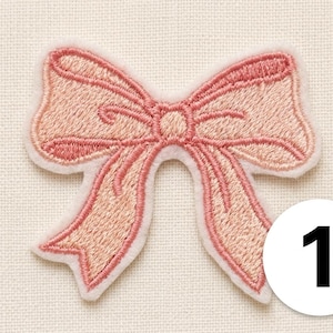 Op de afbeelding: Een lichtroze geborduurde strikpatch met een witte rand en een wit rond label met het zwarte cijfer 1. De strik is voorzien van gedetailleerd stikwerk, wat zorgt voor een gestructureerd uiterlijk met donkerroze draadaccenten langs de randen.