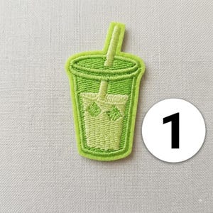 Può includere: Una toppa ricamata a forma di bicchiere con cannuccia, realizzata con fili di colore verde chiaro e verde lime. Il design raffigura un contenitore per bevande con cubetti di ghiaccio visibili all'interno e una cannuccia verticale, accompagnato da un cerchio bianco con il numero 1.