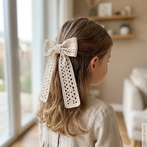May include: A cream-colored crochet hair bow featuring an intricate openwork pattern with two long, rectangular hanging tails. The bow is constructed from yarn and is secured to the hair, displaying a detailed texture and a classic bow shape.