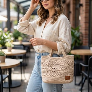 May include: A beige crochet tote bag with two handles and a rectangular brown leather patch featuring the text Toh. The bag has a textured, woven pattern with horizontal rows of varying stitch designs, crafted from a light tan yarn material.