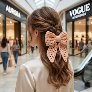 May include: A peach-coloured crochet hair bow with a textured, open-weave pattern. The bow features two large loops and two long, pointed tails, secured to the back of a braided hairstyle. The accessory is crafted from yarn with a visible stitch detail.