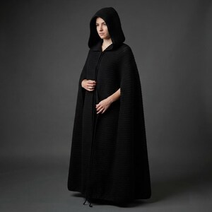 May include: A floor-length black hooded cloak featuring a textured crochet knit pattern. The garment includes a deep hood, an open front design, and long, flowing panels that extend to the floor, providing full coverage in a solid black color.