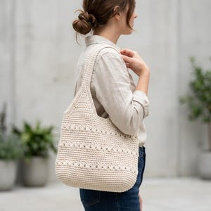 Pode incluir: Uma bolsa de crochê na cor creme com um padrão texturizado de fileiras horizontais de pontos abertos. A bolsa possui uma alça larga de ombro e uma base arredondada e estruturada, feita de fio em um tom neutro de branco off-white.