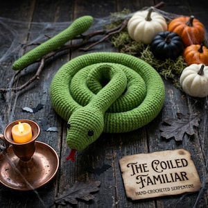 Peut inclure: Un serpent en crochet vert enroulé en cercle avec une langue rouge et des yeux en perles noires. À côté se trouve une plaque en bois avec le texte "The Coiled Familiar" et "Hand-Crocheted Serpentine" imprimé en lettres noires décoratives.