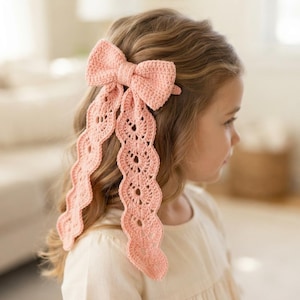 May include: A peach-colored crochet hair bow featuring a central bow knot and two long, wavy, lace-like dangling ribbons with a repeating circular pattern. The accessory is crafted from yarn and is designed to be worn in the hair as a decorative piece.