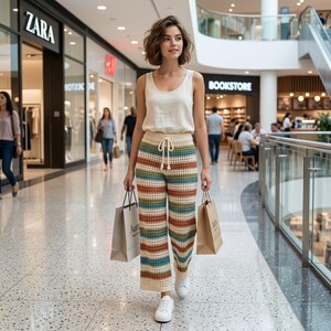 Puede incluir: Una mujer con una camiseta sin mangas color crema y pantalones de crochet a rayas con cintura con cordón camina por un centro comercial. Lleva bolsas de compras y zapatillas blancas. Los pantalones presentan rayas horizontales de varios colores.