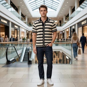 May include: A short-sleeved, button-down knit polo shirt featuring horizontal cream and black stripes, paired with dark blue denim jeans and white sneakers. The shirt has a solid black collar and black trim on the sleeves and hem.