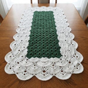 May include: A rectangular crochet table runner featuring a dark green central panel with a repeating floral-like stitch pattern, surrounded by a wide, scalloped white border with a fan-shaped design that extends along all four edges.