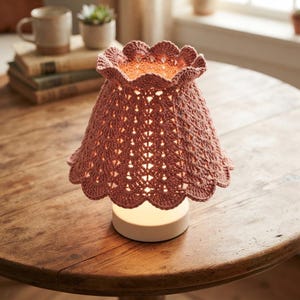 Puede incluir: Una lámpara de mesa con una pantalla de ganchillo en color rosa empolvado, con borde festoneado y un intrincado patrón calado. La pantalla se asienta sobre una base blanca circular sencilla que emite un brillo cálido y suave desde el interior.