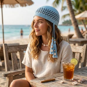 Può includere: Un berretto all'uncinetto azzurro con un motivo a rete traforato sulla calotta e due lunghi laccetti intrecciati laterali. Ogni laccetto è decorato con perline di legno, piccoli accenti turchesi e conchiglie bianche pendenti.