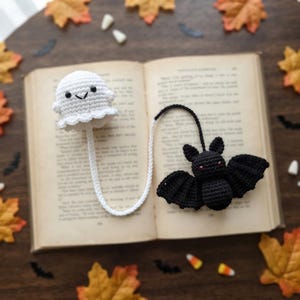 May include: A crochet bookmark featuring a white ghost with black bead eyes and a stitched mouth, connected by a white cord to a black bat with red bead eyes and spread wings. The bookmark is placed on an open book with visible printed text.