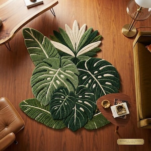 Puede incluir: Una alfombra tejida a mano con forma de grupo de hojas tropicales en varios tonos de verde y crema. La alfombra presenta texturas detalladas que imitan el follaje de monstera y palmera, dispuestas en un patrón orgánico y superpuesto.