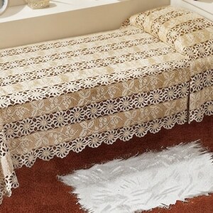 Könnte beinhalten: Eine beige Häkel-Tagesdecke mit einem sich wiederholenden Blumen- und Kreismuster sowie einem gewellten Rand. Das Textil ist über ein Bett drapiert und zeigt komplizierte Durchbrucharbeiten sowie einen einheitlichen hellbraunen Farbton.