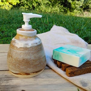 May include: A ceramic soap dispenser with a white pump and a brown, ribbed, tapered body featuring a white glazed top. Beside it sits a square bar of white soap with teal swirls resting on a dark brown wooden soap dish atop a white cloth.