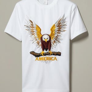 Può includere: Una t-shirt bianca a maniche corte con un grande ricamo dettagliato di un'aquila calva con le ali spiegate appollaiata su un ramo. L'aquila ha piume marroni, bianche e gialle. La parola AMERICA è ricamata in giallo nella parte inferiore.