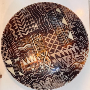 May include: A round ceramic bowl featuring a dark brown and cream-colored surface with a complex, repeating geometric pattern. The design includes various sections of zigzags, cross-hatching, wavy lines, and triangular shapes across the interior.