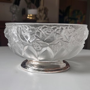 May include: A clear glass bowl featuring a frosted relief pattern of blooming roses and leaves around the exterior. The bowl sits on a circular, polished silver-toned metal base that provides a reflective contrast to the textured glass upper section.