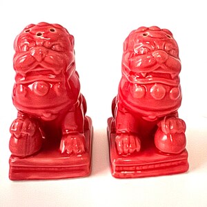 May include: A pair of bright red ceramic Foo Dog salt and pepper shakers. Each figure features a stylized lion-like face with a mane, sitting on a rectangular base. The top of each head has small holes for dispensing salt and pepper seasoning.