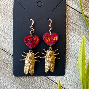 May include: A pair of dangling earrings featuring a heart-shaped top with a red and pink glitter design, connected to a gold-colored cicada charm. The earrings hang from a gold-colored hook. The earrings are displayed on a black card.