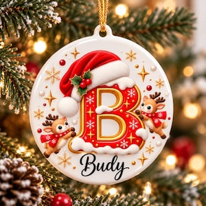 Puede incluir: Un adorno navideño redondo de cerámica blanca con una letra B grande en rojo y dorado, coronada con un gorro de Papá Noel y acebo. Dos renos de dibujos animados con bufandas rojas flanquean la letra. El nombre Budy aparece en negro.