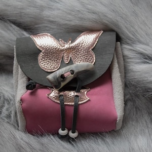May include: A small pouch featuring a dark gray flap adorned with a metallic rose gold butterfly applique. The body is a deep pink color with light gray side panels. A gray toggle closure secures the flap, and two black cords with white beads hang from the front.