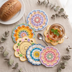 May include: A collection of six round, multi-colored crochet coasters featuring intricate mandala-style patterns in shades of yellow, pink, blue, purple, and white. The coasters are arranged in a cluster, with one supporting a glass of iced tea.