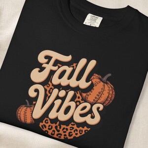 Puede incluir: Una camiseta negra con el texto Fall Vibes en una fuente retro de color tostado claro. El diseño incluye dos calabazas naranjas y un elemento circular de fondo con un estampado de leopardo marrón, centrado en la parte frontal de la prenda.