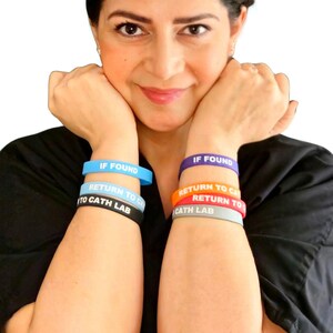 Puede incluir: Un conjunto de seis pulseras de silicona que se llevan en los antebrazos. El brazo izquierdo tiene pulseras azul claro, blancas y negras, mientras que el derecho tiene pulseras moradas, naranjas y grises. Cada una lleva el texto IF FOUND RETURN TO CATH LAB.