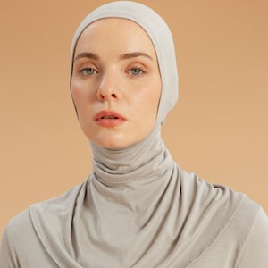 May include: A light grey jersey hijab cap that covers the head and neck, featuring a smooth, fitted fabric construction that drapes around the collarbone. The material is a soft, solid-coloured knit textile that provides full coverage.