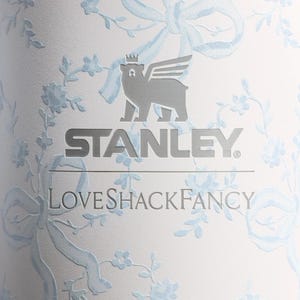Puede incluir: Un vaso blanco con un patr&oacute;n de flores y cintas en azul claro. La superficie muestra el logotipo plateado de Stanley con un icono de oso alado encima, seguido del texto LoveShackFancy en una fuente serif plateada centrado debajo.