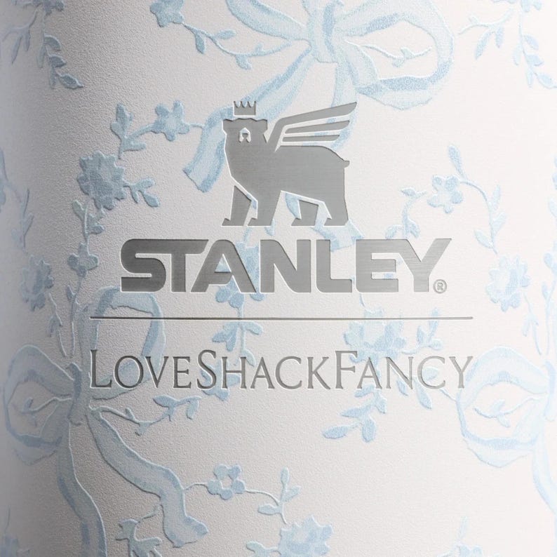 Puede incluir: Un vaso blanco con un patr&oacute;n de flores y cintas en azul claro. La superficie muestra el logotipo plateado de Stanley con un icono de oso alado encima, seguido del texto LoveShackFancy en una fuente serif plateada centrado debajo.
