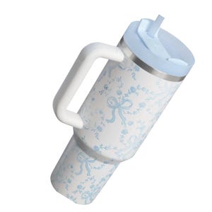 Puede incluir: Un vaso de acero inoxidable blanco con un patr&oacute;n de flores y lazos en azul claro, que cuenta con un asa lateral blanca resistente, una tapa azul claro y una pajita a juego. El recipiente cil&iacute;ndrico tiene una base c&oacute;nica y una tapa plana.