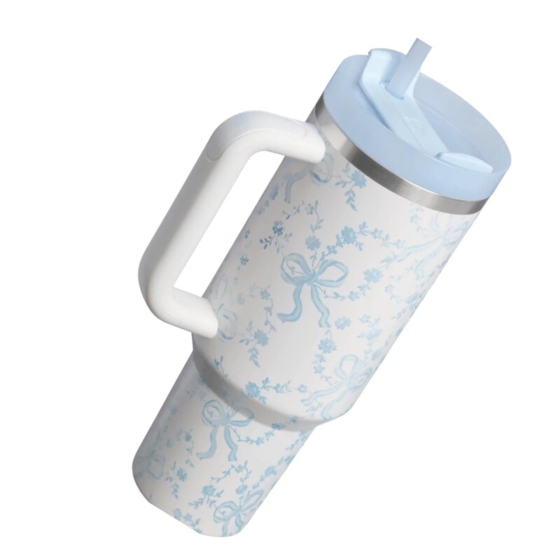 Puede incluir: Un vaso de acero inoxidable blanco con un patr&oacute;n de flores y lazos en azul claro, que cuenta con un asa lateral blanca resistente, una tapa azul claro y una pajita a juego. El recipiente cil&iacute;ndrico tiene una base c&oacute;nica y una tapa plana.