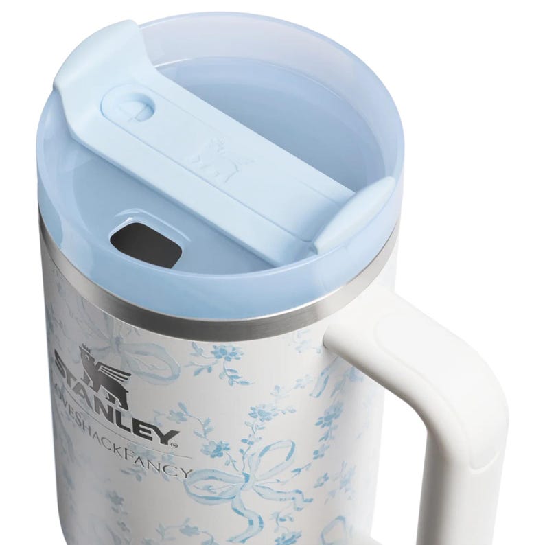Puede incluir: Un vaso de acero inoxidable blanco con un patr&oacute;n de flores y lazos en azul claro. La taza cuenta con una tapa azul claro con cierre deslizante y un asa blanca. El lateral muestra los logotipos negros de Stanley y LoveShackFancy impresos.