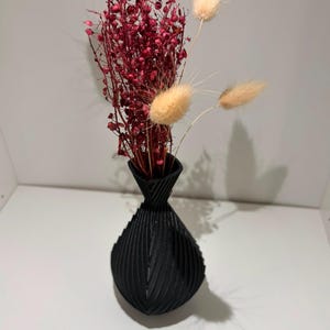 May include: A matte black vase with a ribbed, geometric texture and a narrow neck. The vase contains a bouquet of dried flowers, featuring vibrant red, branching stems and three soft, beige, oval-shaped bunny tail grass plumes.