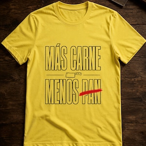 Puede incluir: Una camiseta amarilla de manga corta con el texto negro MÁS CARNE MENOS PAN, un pequeño gráfico de un cuchillo de carnicero entre las líneas y una línea horizontal roja que tacha la palabra PAN en la línea inferior del texto.