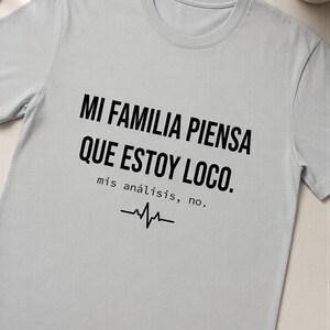 Op de afbeelding: Een lichtgrijs t-shirt met korte mouwen en zwarte tekst die luidt MI FAMILIA PIENSA QUE ESTOY LOCO. mis análisis, no. met daaronder een zwarte afbeelding van een hartslaglijn. Het t-shirt heeft een ronde hals en een standaard pasvorm.